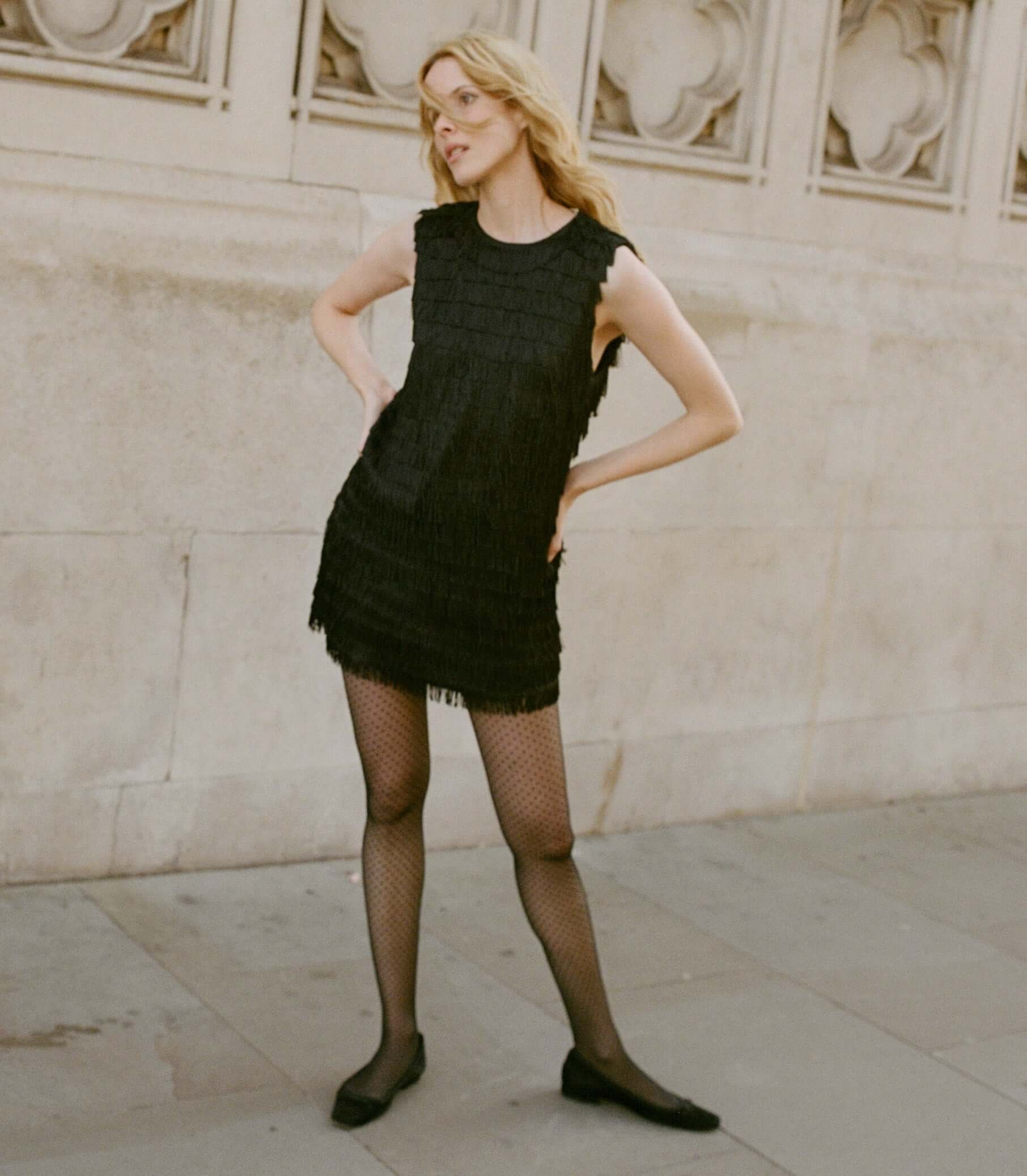 A model posing in a fringed black dress and fishnet stockings, showcasing stylish tops.